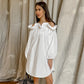 A woman models a Kaelen white off-shoulder cotton mini dress with dramatic ruffled neckline and voluminous balloon sleeves, standing against a textured neutral backdrop.