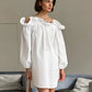 A white Kaelen off-shoulder cotton mini dress with voluminous balloon sleeves is showcased on a model, styled in a chic, relaxed indoor setting with soft lighting.