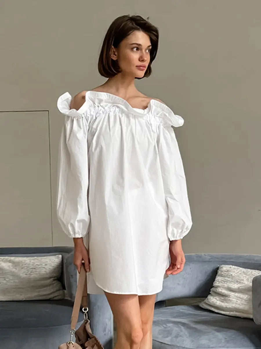 A white Kaelen off-shoulder cotton mini dress with voluminous balloon sleeves is showcased on a model, styled in a chic, relaxed indoor setting with soft lighting.