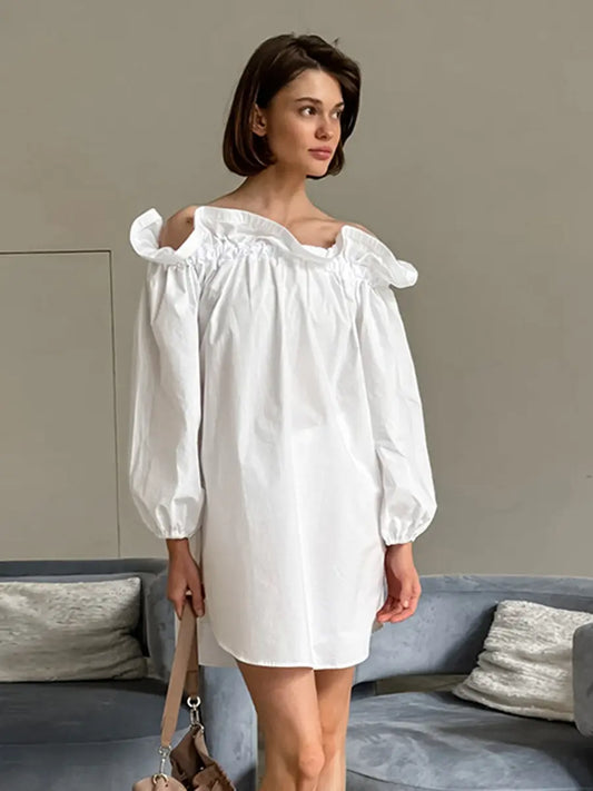 A white Kaelen off-shoulder cotton mini dress with voluminous balloon sleeves is showcased on a model, styled in a chic, relaxed indoor setting with soft lighting.