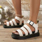White Kaelen platform gladiator sandals with a chunky black sole and strappy faux leather upper, worn on feet against a rustic wooden floor. Features adjustable ankle buckle.