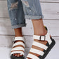 Woman's feet wearing white Kaelen platform gladiator sandals with black buckles and yellow stitching detail, paired with distressed denim jeans against a wood paneled background.