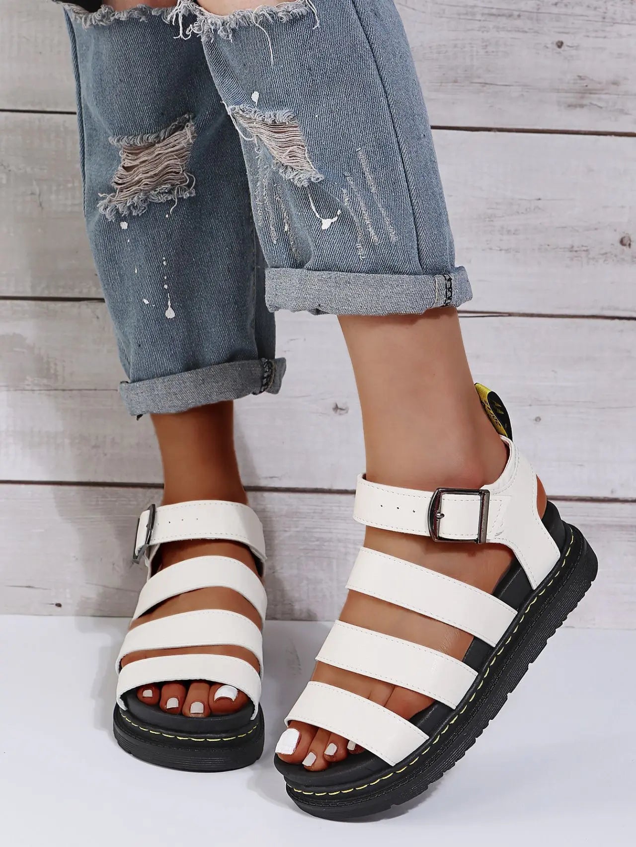 Woman's feet wearing white Kaelen platform gladiator sandals with black buckles and yellow stitching detail, paired with distressed denim jeans against a wood paneled background.