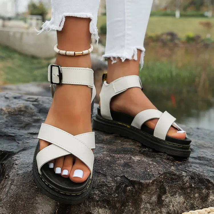 White Kaelen platform sandals with criss-cross straps and a buckle ankle strap, worn with ripped white jeans on a rocky, natural outdoor background.