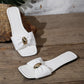 Pair of Kaelen white woven flat slide sandals with square toes and oval gold buckles, displayed on a rustic wooden surface.
