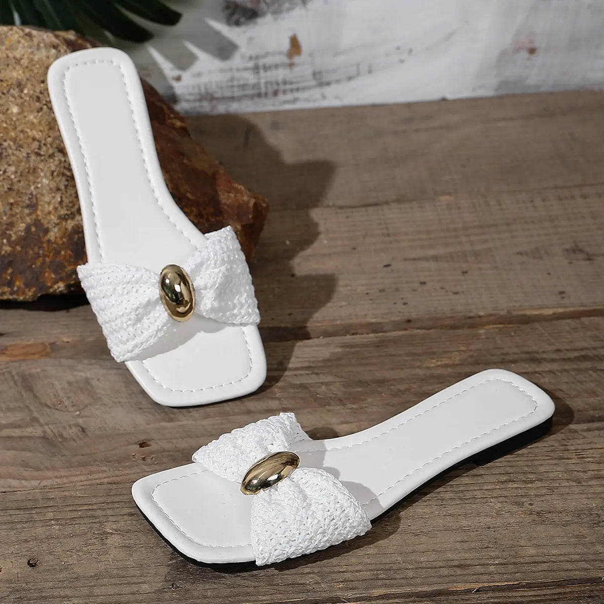 Pair of Kaelen white woven flat slide sandals with square toes and oval gold buckles, displayed on a rustic wooden surface.