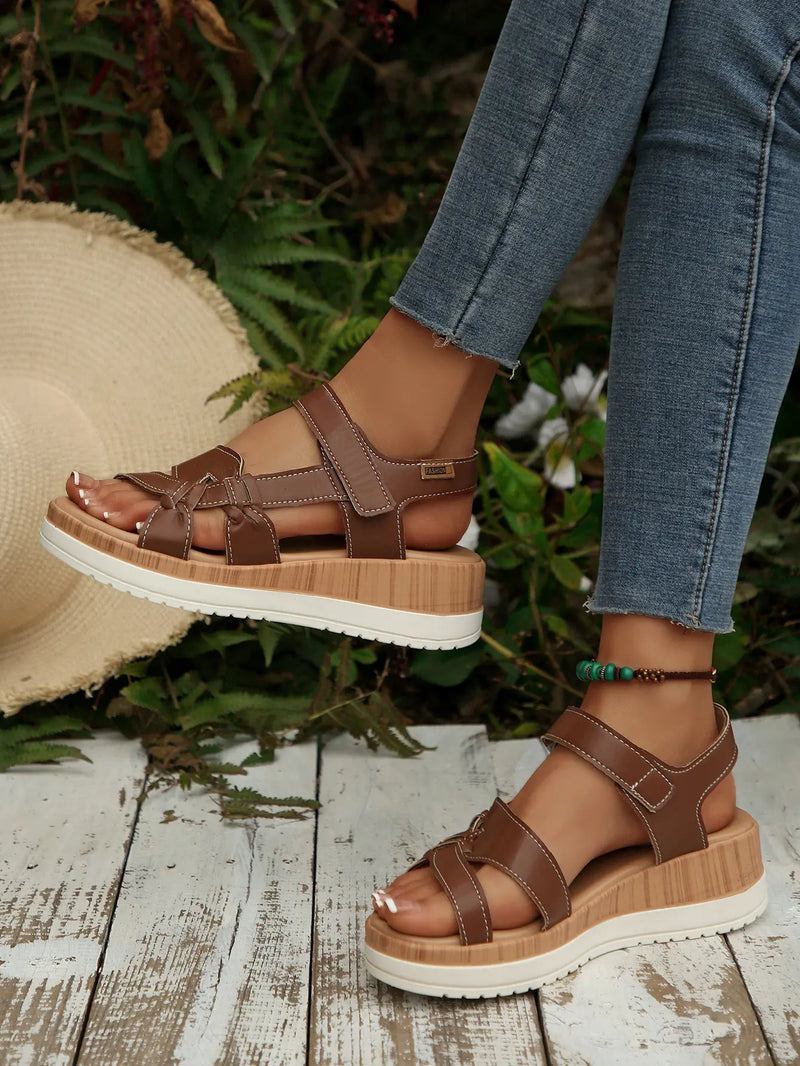Brown Kaelen women's flat sandals with multi-strap design and knot detail, worn on feet with jeans and a straw hat nearby. Features hook-and-loop closure and a wood-grain effect platform sole.