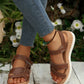 Brown faux leather Kaelen women's flat sandals with adjustable hook-and-loop straps and a decorative knot detail, shown on a person's feet with jeans in a natural outdoor setting.