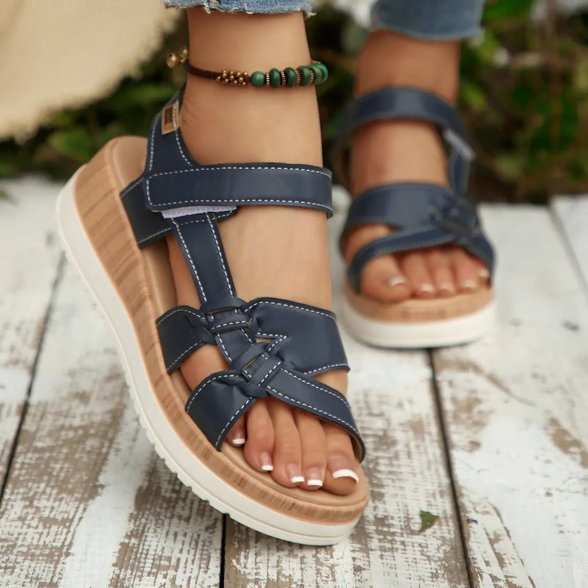 Kaelen women's dark blue flat sandals with faux leather multi-strap design and decorative knot detail, worn on feet with French tip pedicure, set on a white wooden plank with a blurred green background.