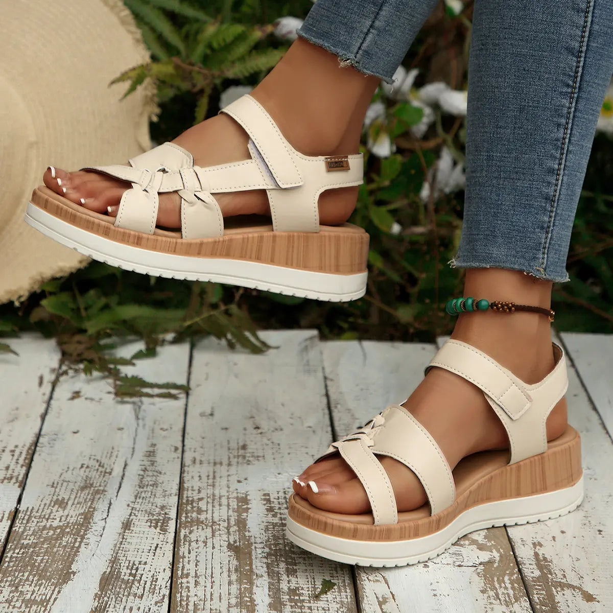 Kaelen women's flat sandals in beige faux leather, featuring a knot detail on the forefoot strap and adjustable hook-and-loop closure, worn on feet over a distressed wooden surface with natural foliage in the background.