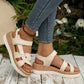 Model wearing beige Kaelen women's flat sandals with faux leather straps, a decorative knot detail on the forefoot, and a wood-grain effect platform sole in a natural outdoor setting.