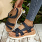 Close-up of navy blue Kaelen women's flat sandals worn on a foot, featuring multi-straps with knot detail, adjustable hook-and-loop closure, and a wood-grain effect platform sole against a natural, blurred outdoor backdrop.