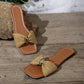 Kaelen woven flat slide sandals in natural raffia with a gold oval buckle, featuring a square open toe and laid on a rustic wooden surface.