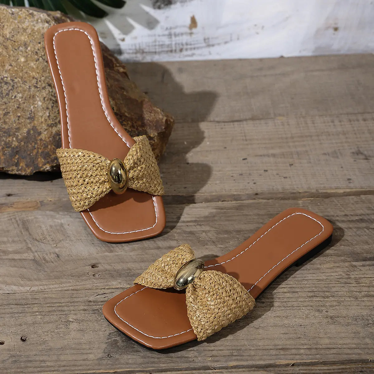 Kaelen woven flat slide sandals in natural raffia with a gold oval buckle, featuring a square open toe and laid on a rustic wooden surface.
