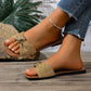 Kaelen woven flat slide sandals with a gold oval buckle detail, shown on a woman's feet paired with distressed denim jeans on a rustic wooden background.