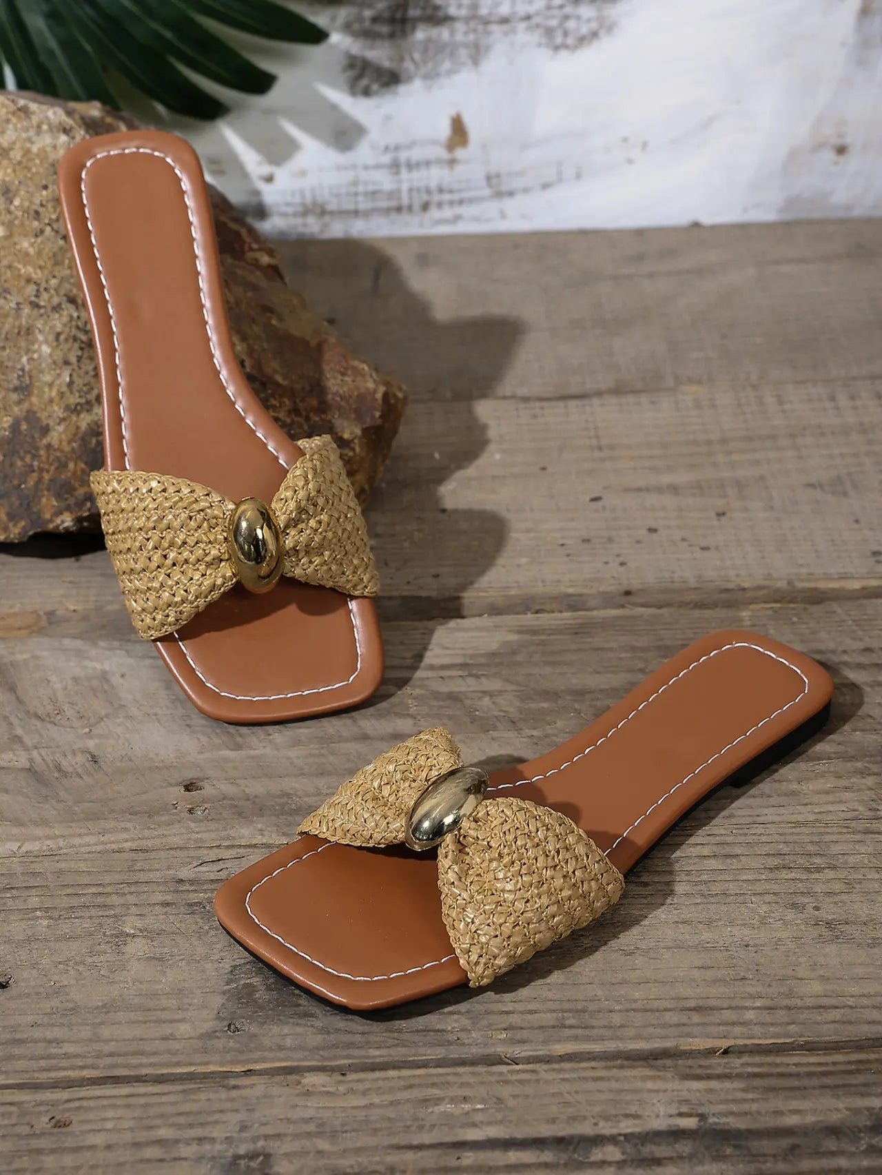 Kaelen woven flat slide sandals in natural raffia with a gold oval buckle, shown from a three-quarter angle on a wooden surface with natural stone elements.