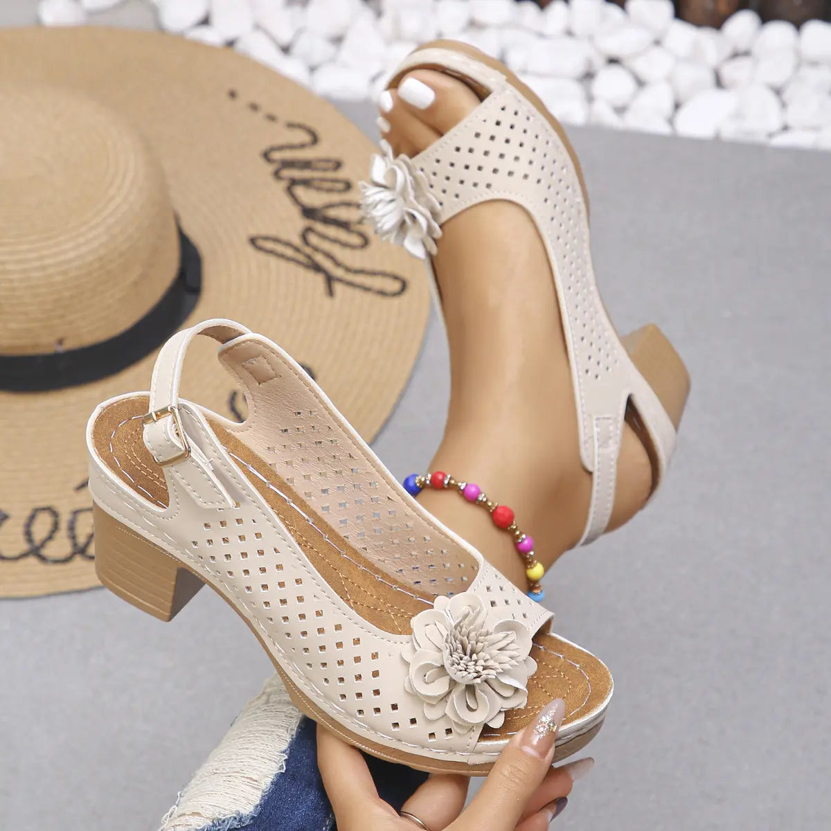 Kaelia beige peep-toe chunky heel sandals adorned with layered floral embellishments and intricate square perforations on the upper, shown on a model's foot alongside a straw hat.