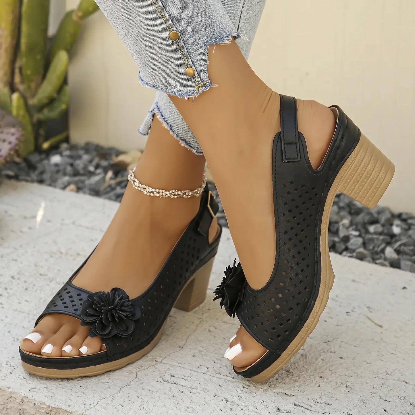 Kaelia black chunky heel peep-toe sandals adorned with a floral embellishment and perforated square pattern, shown on a model's feet with a slingback strap and ankle strap detail.
