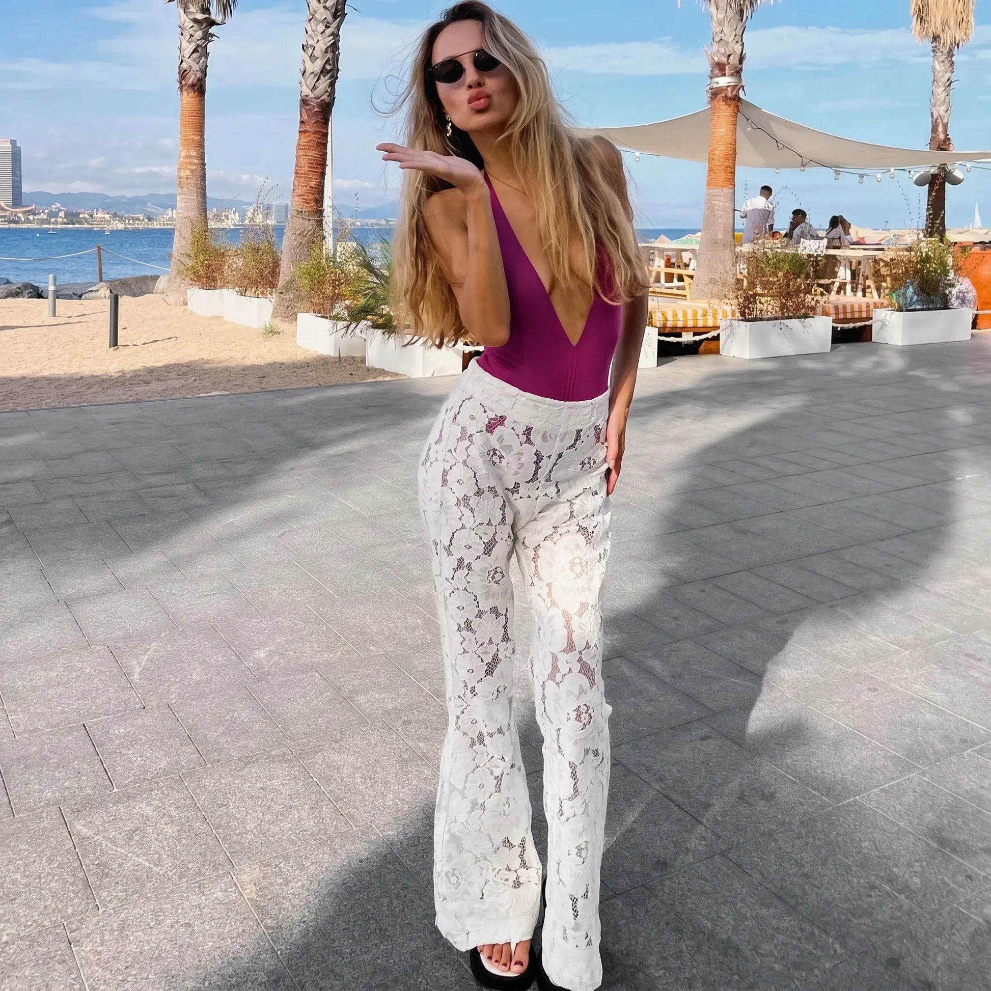 A woman models elegant white lace flared trousers with a high waist and bohemian design, styled for a resort setting on a sunny promenade.