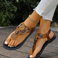 A woman's feet wearing Kaelin bohemian brown flat sandals adorned with multicolor beads and golden mandala charms, styled with white jeans and an ankle bracelet on a wooden deck.