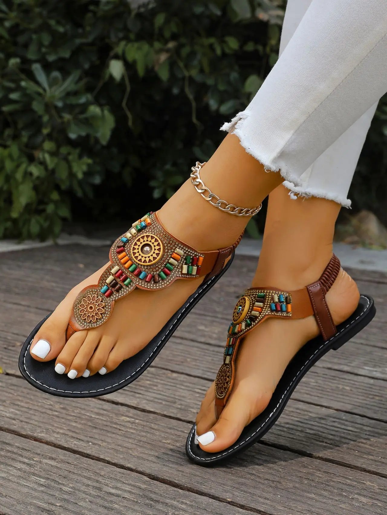 A woman's feet wearing Kaelin bohemian brown flat sandals adorned with multicolor beads and golden mandala charms, styled with white jeans and an ankle bracelet on a wooden deck.