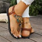 Close-up of Kaelin artisanal bohemian flat sandals with multicolor beadwork and golden mandala charms, worn on a wooden deck with white jeans. Features T-strap, elastic ankle strap, and intricate detailing.