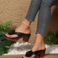 Kaelis black floral platform sandals worn on a model's feet, styled with distressed denim jeans and a pearl anklet, set against a rustic stone and wood background.