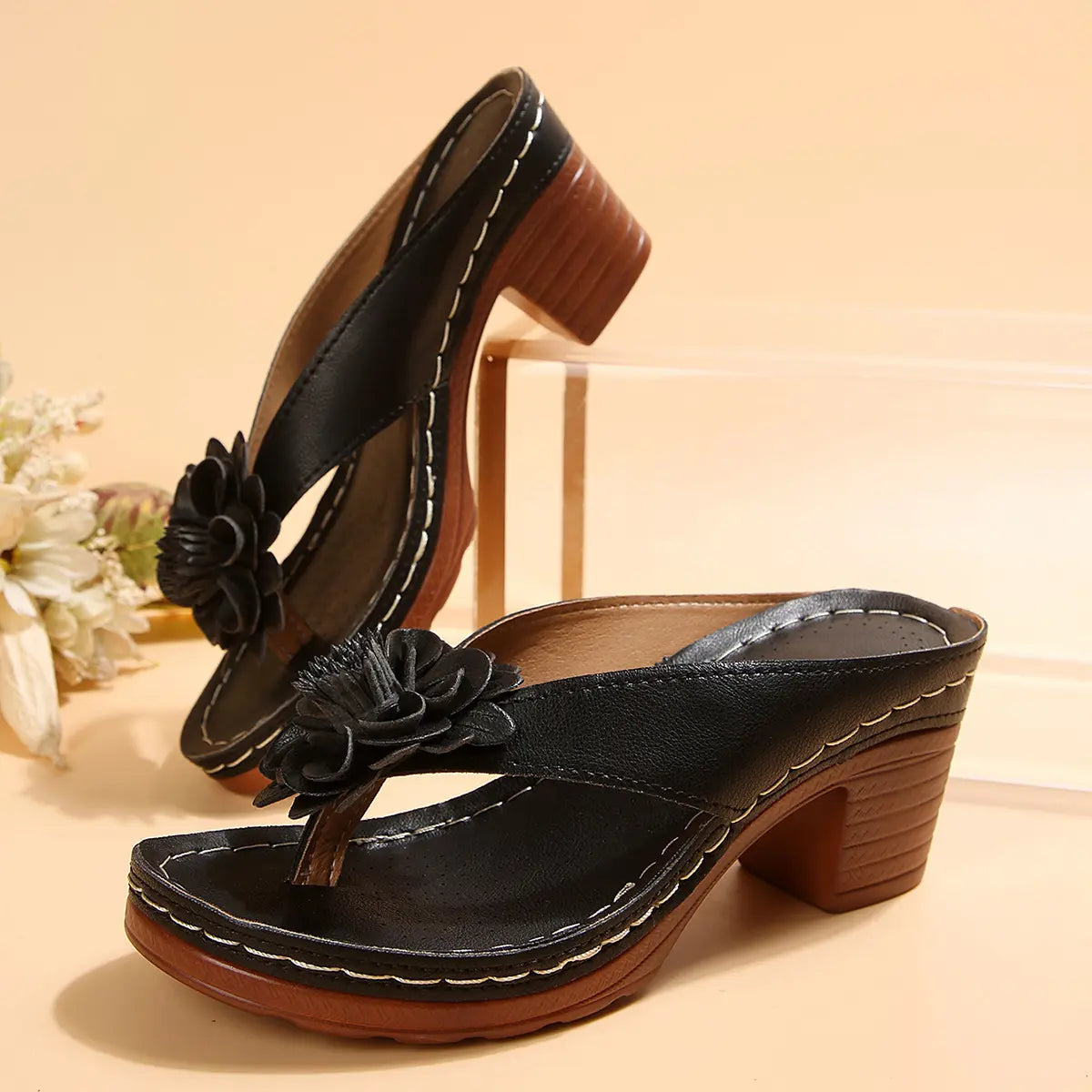 Kaelis black floral platform sandals featuring a wide faux leather strap adorned with a 3D fabric flower, a contoured footbed, and a chunky wood-effect block heel. Shown from a three-quarter front view.
