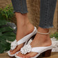 Kaelis white floral platform sandals worn by a model with distressed denim jeans, showcasing the chunky block heel and 3D flower embellishment on a rustic wooden and pebble-filled backdrop.