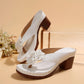 White Kaelis floral platform sandals featuring a large 3D flower embellishment on a wide strap, a chunky wood-effect block heel, and a layered sole. Presented in a studio setting with decorative elements.