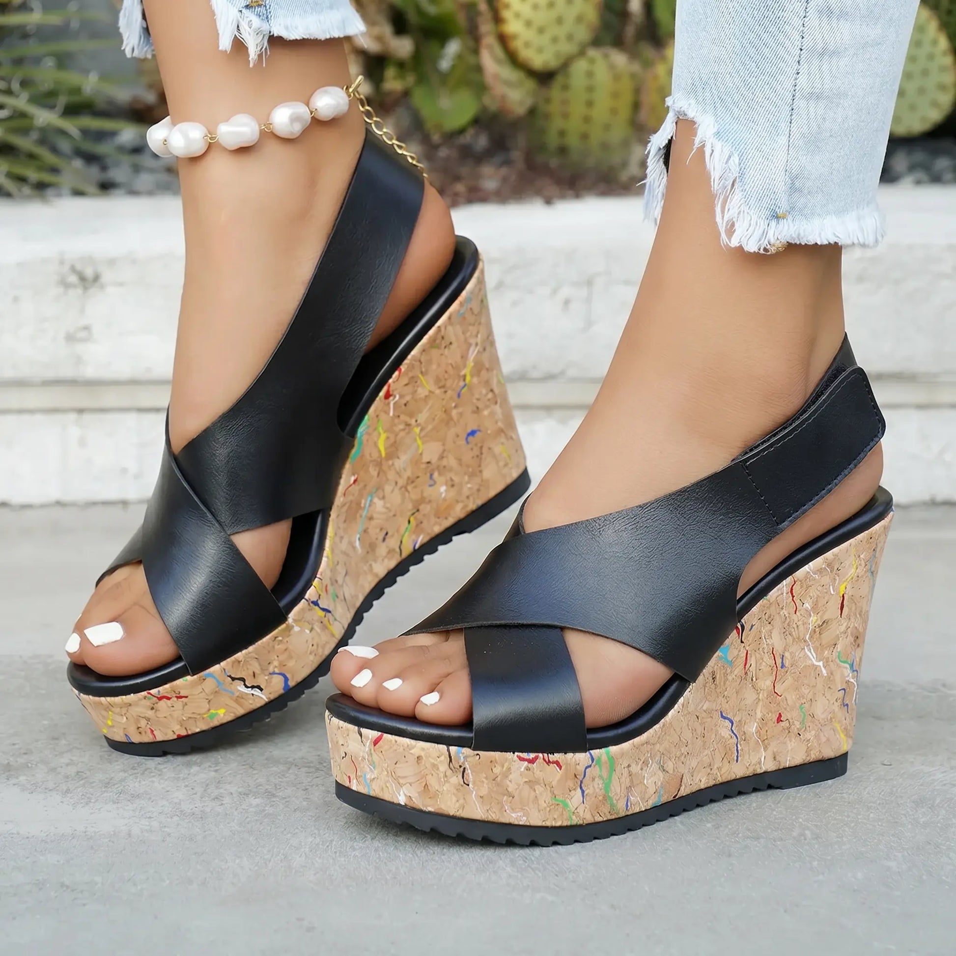 Kaelo black faux leather criss-cross strap wedge sandals with a multi-color flecked cork platform and heel, shown on feet with distressed jeans and a pearl ankle bracelet.