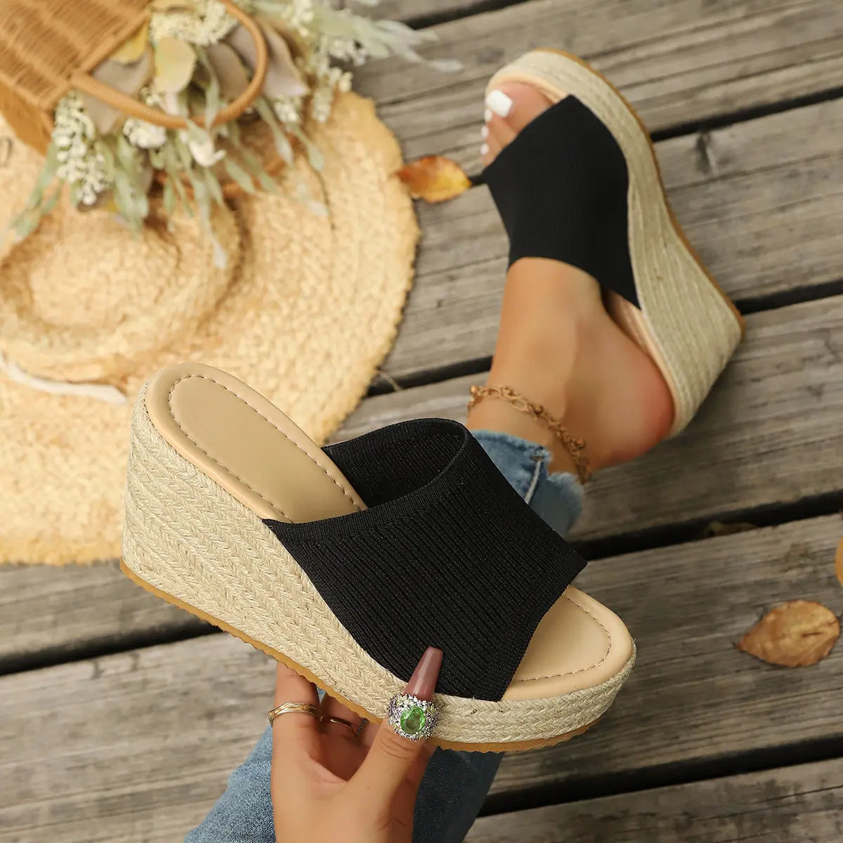 Black knitted espadrille wedge sandals with an open toe and platform sole, shown on a woman's feet styled on a rustic wooden boardwalk with a straw hat.