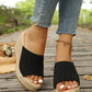 Black knitted espadrille wedge sandals with a chunky platform and open toe, shown on feet with frayed denim jeans on a wooden boardwalk.