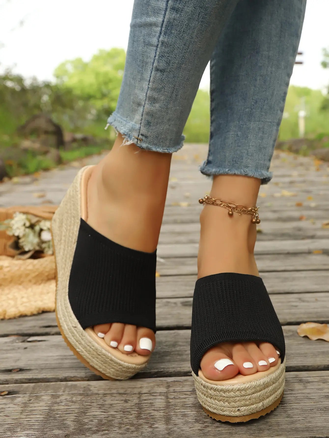 Black knitted espadrille wedge sandals with a chunky platform and open toe, shown on feet with frayed denim jeans on a wooden boardwalk.
