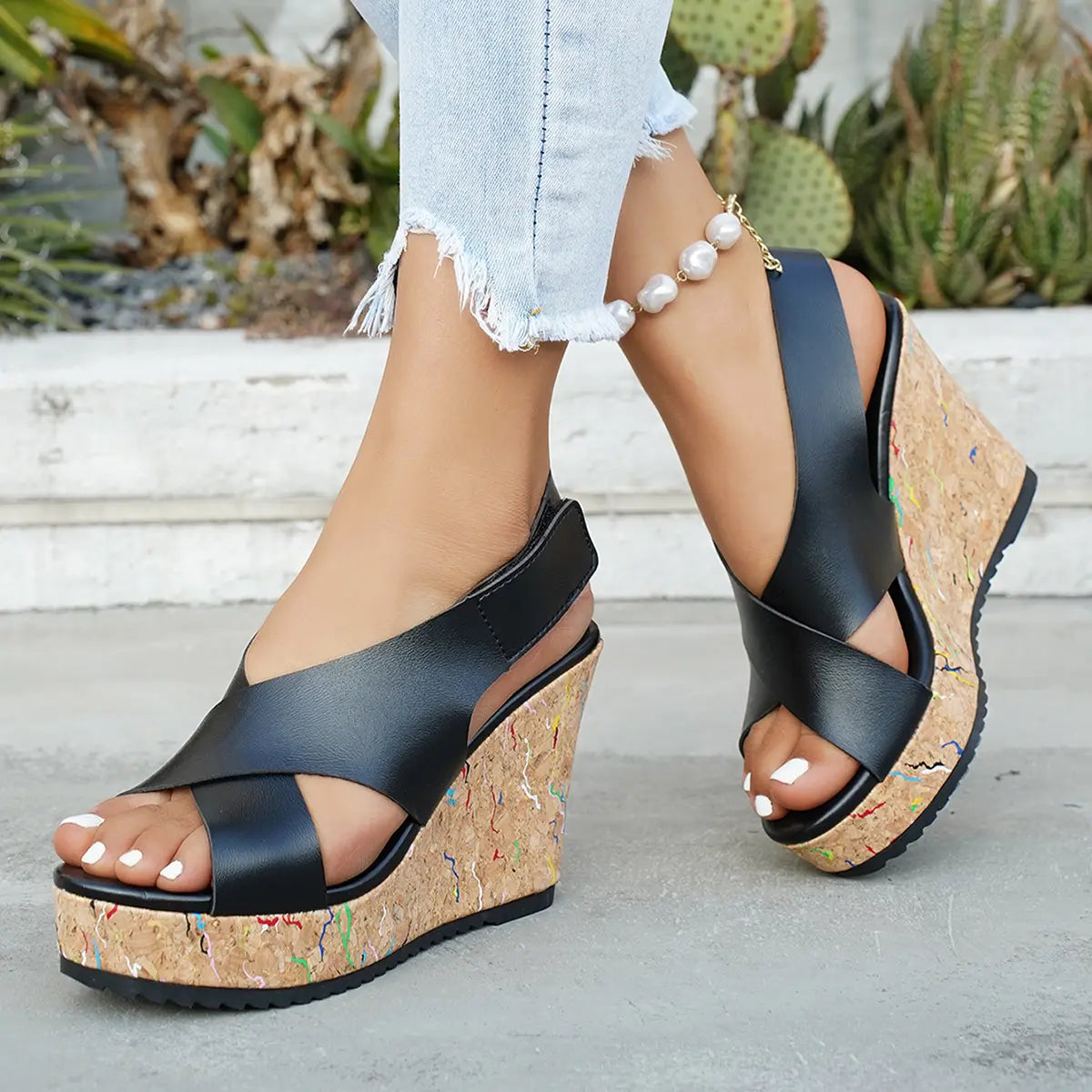 Kaelo black faux leather criss-cross strap wedge sandals with a multi-colored speckled cork platform and heel, worn by a model on a concrete path with cacti in the background.
