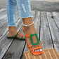 A pair of Kaelo color block flat slide sandals, featuring vibrant orange straps with a green geometric accent on one sandal, worn by a person in jeans on a wooden deck.