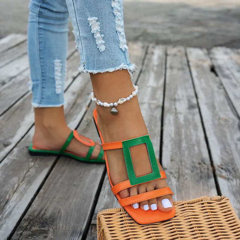 A pair of Kaelo color block flat slide sandals, featuring vibrant orange straps with a green geometric accent on one sandal, worn by a person in jeans on a wooden deck.