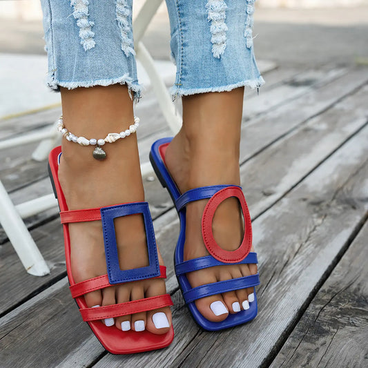 Kaelo women's asymmetrical color-block flat slide sandals in red and blue, featuring geometric and circular cut-outs, shown on feet with ripped denim jeans.