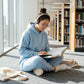 Kaelo premium cropped hoodie and jogger lounge set in light blue, worn by a woman sitting on a library floor reading with headphones.