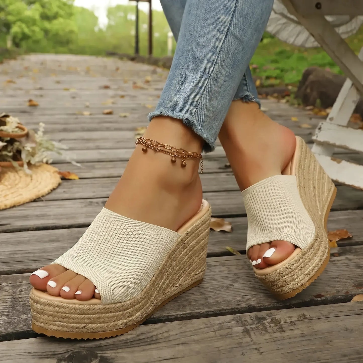 Women's Kaelo Natural Linen knitted espadrille wedge sandals are shown on feet, styled with blue denim jeans and an anklet, resting on a rustic wooden boardwalk with scattered leaves.