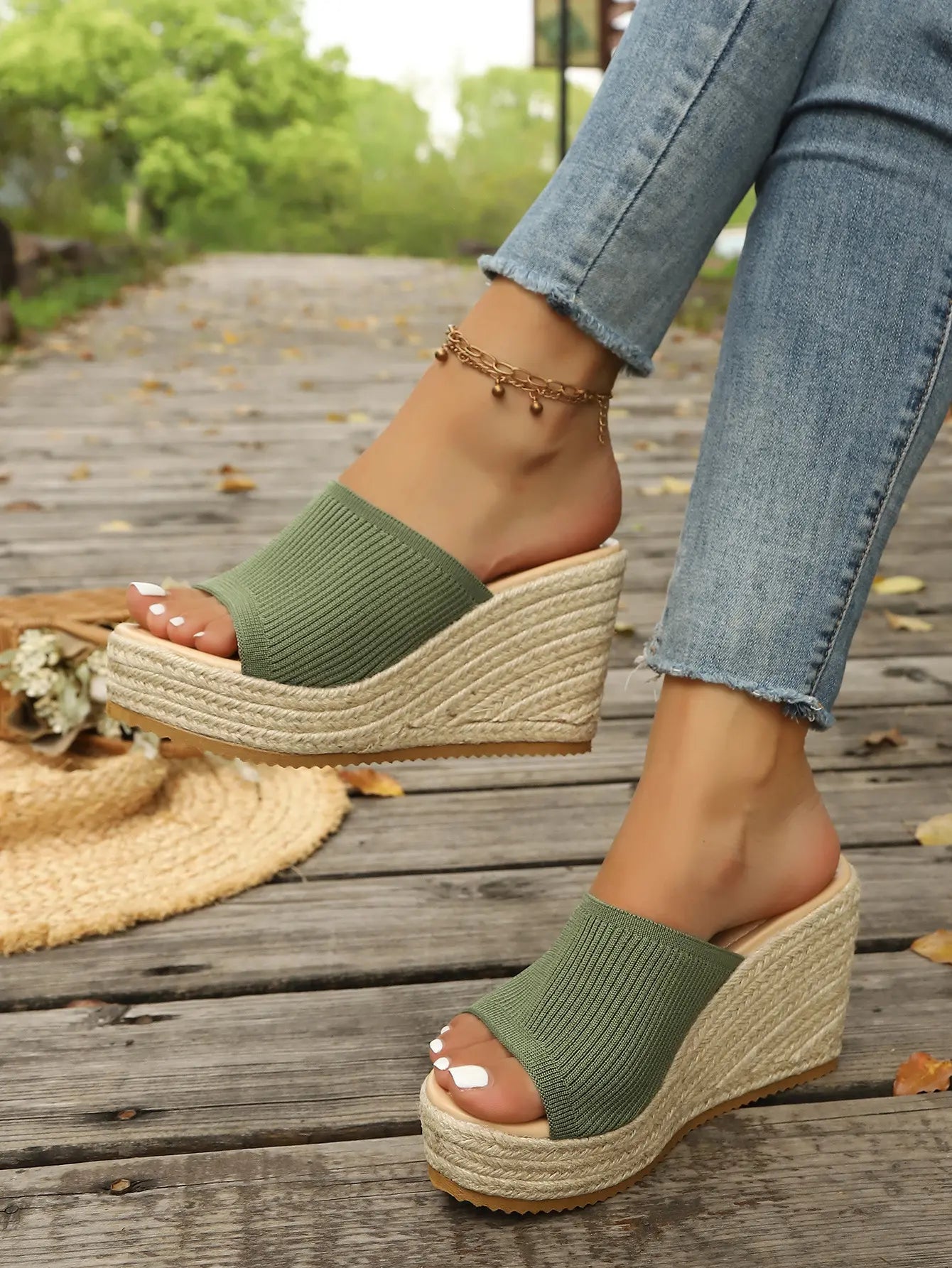 Olive green knitted espadrille wedge sandals with a thick jute-wrapped platform sole are showcased on a woman's feet, styled with casual blue jeans on a wooden boardwalk.