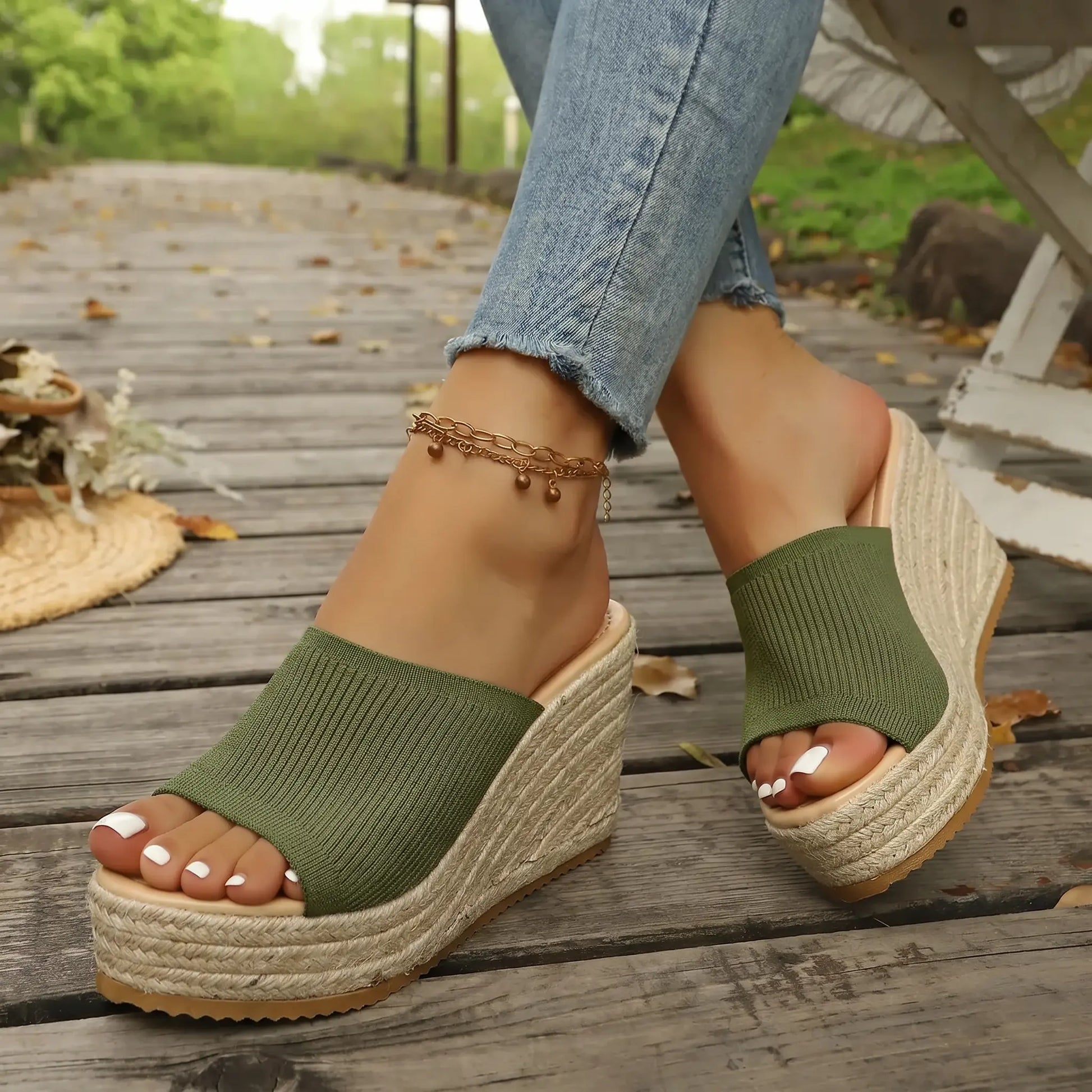 Kaelo olive green knitted espadrille wedge sandals on a woman's feet, styled with blue jeans on a wooden boardwalk with dried leaves, showcasing the open-toe design and jute-wrapped platform sole.