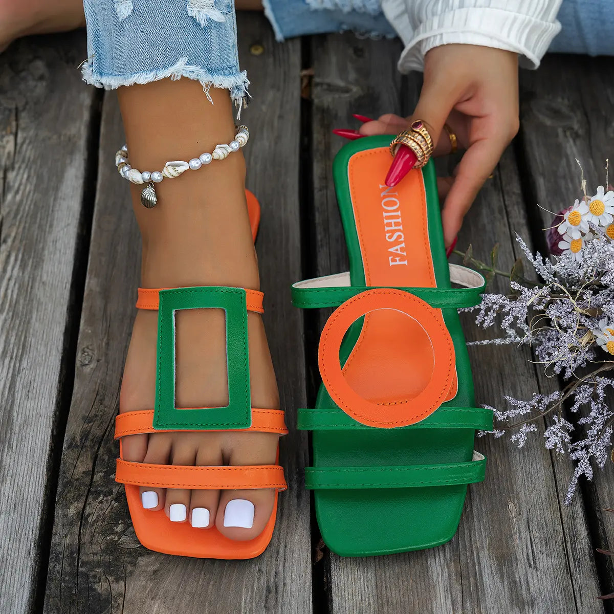 Kaelo flat slide sandals in vibrant orange and green color-block design, featuring geometric and circular cut-outs on an open square toe. Shown on feet with white toenails on a wooden surface.