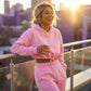 Kaelo pink cropped hoodie and jogger set worn by a smiling woman on a balcony, showing the adjustable toggle waist and relaxed fleece fit.