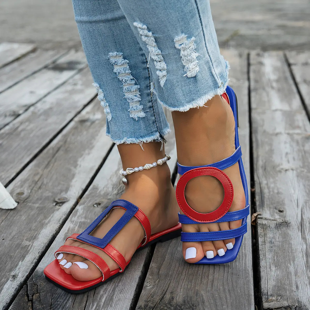 Kaelo women's red and blue asymmetrical color block flat slide sandals worn with distressed jeans on a wooden deck.