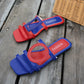 Kaelo vibrant red and blue asymmetrical color-block flat slide sandals with geometric cut-out designs and 'FASHION' printed on the footbed, displayed on a wooden surface.