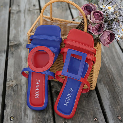 Bold red and blue Kaelo flat slide sandals with asymmetrical color-blocking and geometric accents, presented on a rustic wooden surface with a woven basket and floral arrangement.