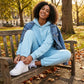Kaelo women's cropped hoodie and jogger set in light blue — lifestyle view sitting on a park bench styled with a denim jacket.