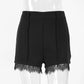 Black high-waisted tailored shorts featuring delicate floral lace trim on the hem and sharp front pleats, presented from a front view on a mannequin.