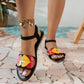 Black flat sandals for women featuring vibrant multi-colored heart appliques on the front strap and a secure black elasticated ankle strap with a buckle, shown on a person's feet against a scenic tiled and water background.
